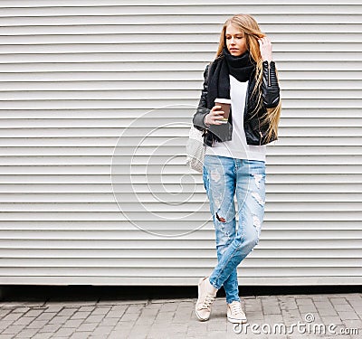 Portrait of a beautiful young blonde girl with long hair posing on a street with coffee and a backpack. Outdoor, warm color. Stock Photo