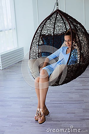 Portrait of beautiful young brunette woman in blue dress with make up and ponytail, sitting in large chair Stock Photo