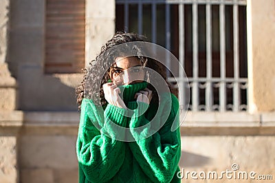 Portrait of beautiful young brunette woman with curly hair and green woollen coat covering her face with the collar of the coat Stock Photo
