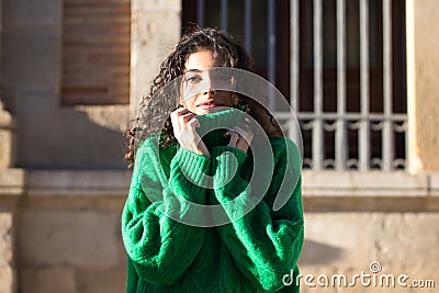 Portrait of beautiful young brunette woman with curly hair and green woollen coat covering her face with the collar of the coat Stock Photo