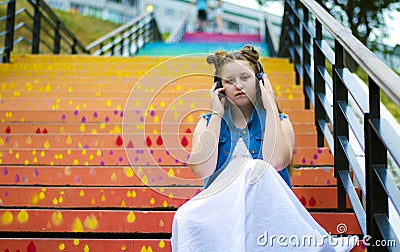 Portrait of a beautiful, young girl who sits on the stairs and listens to music on headphones, in the street, in summer Stock Photo