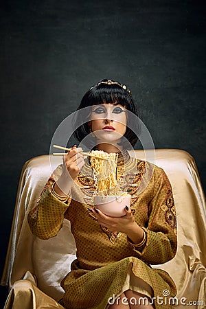 Portrait of beautiful young woman in image of egyptian queen, Cleopatra eating instant noodles against dark vintage Stock Photo