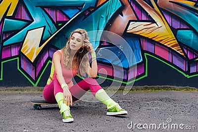 Portrait of beautiful young woman in pink bodysuit sitting on skateboard near wall with graffiti Editorial Stock Photo