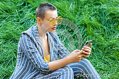 Portrait of beautiful young woman with short hair in casual blue striped suit, yellow glasses sitting on green grass holding her Stock Photo