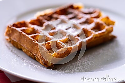 A portrait of a belgian delicious tasty waffle with powder sugar as a topping on it, lying on a white plate on a red table cloth Stock Photo