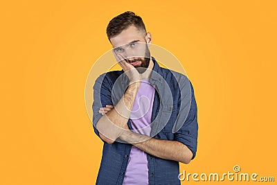 Portrait of bored Caucasian guy leaning on palm and looking at camera on orange studio background Stock Photo