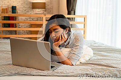 Portrait of bored young Asian girl lying down on bed while working from home in bedroom. Female freelancer working on Stock Photo