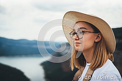 Portrait of brooding young girl with blonde hair in straw summer hat and hipster glasses on backdrop river natura mountain land Stock Photo