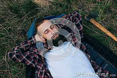 Portrait brutal bearded and moustached woodcutter hipster Gypsy man in the forest with ax. Man lying on the grass and dreams Stock Photo