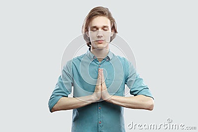 Portrait of calm handsome long haired blonde young man in blue casual shirt standing in yoga pose and meditating, and try to relax Stock Photo