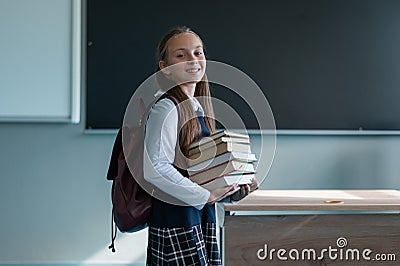 Portrait of a caucasian schoolgirl with a backpack. The girl is holding a stack of textbooks in the classroom. Stock Photo