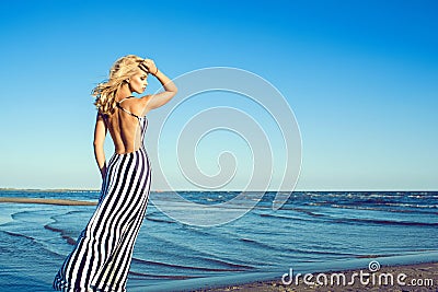 Portrait of charming blond long-haired woman in long black and white striped dress with naked back walking along the seaside Stock Photo