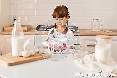 Portrait of charming cute dark haired female kid with pigtails sitting at table and using smart phone, finding recipe in the Stock Photo