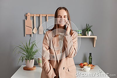 Portrait of cheerful joyful brown haired woman wearing beige suit standing near table on kitchen at home, looking at camera, Stock Photo
