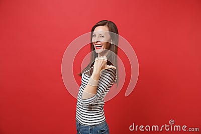 Portrait of cheerful laughing young woman in striped clothes pointing thumb behind her back isolated on bright red wall Stock Photo