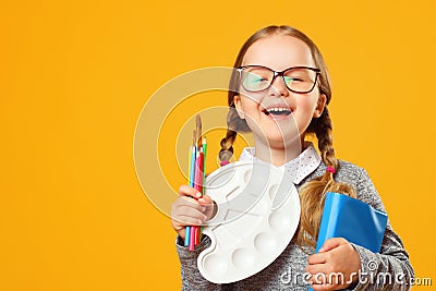 Portrait of a cheerful little child girl on a yellow background. Schoolgirl holds a book, pencils, brushes and a palette. Stock Photo
