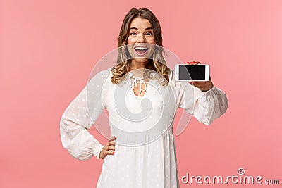 Portrait of cheerful, upbeat attractive blond caucasian girl in white dress, showing smartphone display, hold mobile Stock Photo