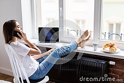 Portrait of cheerful young woman sittingon chair with legs on wooden windowsill with breakfast talking on the phone. Stock Photo