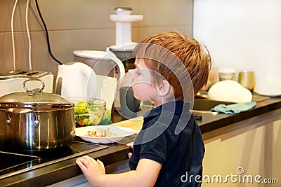 Portrait of child in kitchen. Cute little boy, playing in kitchen. Young kid is hungry, looks at and checks is it finished lunch. Stock Photo