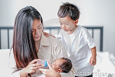 Portrait and close up young Asian mother give milk to her newborn baby via bottle with her little boy look like interest to help Stock Photo