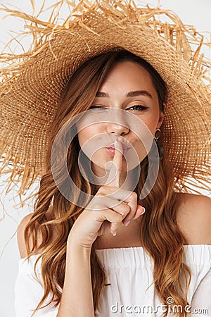 Portrait closeup of european charming woman 20s wearing big straw hat showing finger at lips to keep secret, isolated over white Stock Photo