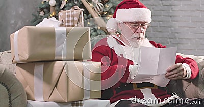 Portrait of concentrated old Santa Claus reading letter. Mature man with white beard in eyeglasses choosing present for. Child. Decorated Christmas tree stock footage Portrait of concentrated old Santa Claus reading letter. Mature man with white beard in eyeglasses choosing present for. Child. Decorated Christmas tree stock footage