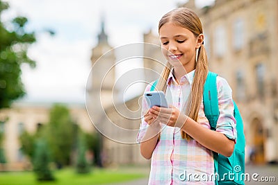 Portrait of concentrated small girl using device holding rucksack backpack wearing checkered plaid t-shirt standing Stock Photo