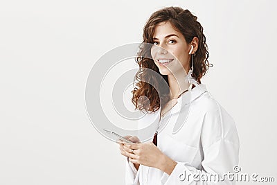 Portrait of confident charming successful woman holding smartphone in hands and wireless earphone in ear, standing half Stock Photo