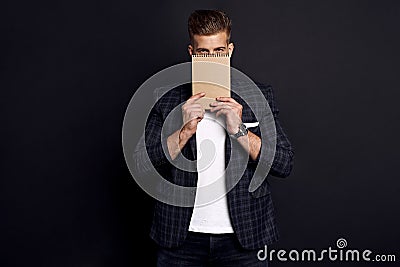 Portrait of confident concentrated self-assured handsome bearded guy covers his face with a notebook, isolated on black background Stock Photo