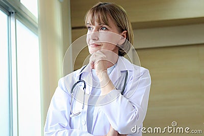 Portrait of confident female doctor in workwear with stethoscope thinking and taking decisions with hand on chin, looking through Stock Photo