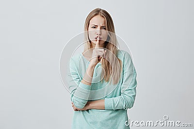 Portrait of confident female with long blonde hair wears blue clothes, shows silence sign, asks to keep private Stock Photo