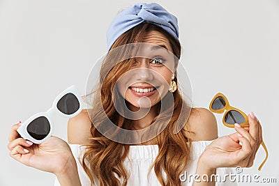 Portrait of confused young woman 20s hesitating while holding two pairs of sunglasses, isolated over white background Stock Photo