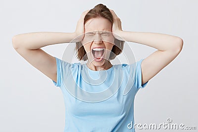 Portrait of crazy hysterical young woman in blue tshirt with closed eyes and hands on head looks stressed and screaming isolated Stock Photo