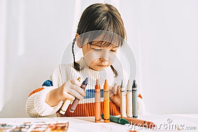 Portrait of creative cute little girl playing with oil pencils, sitting at white desk at home. Pretty preschool kid draws Stock Photo