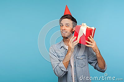 Portrait of curious man with funny party hat holding gift box near ear and listening, guessing present Stock Photo