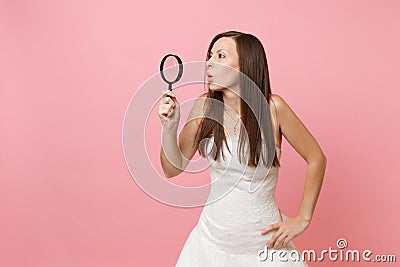 Portrait of curious shocked bride woman in white wedding dress looking aside through magnifying glass on pastel Stock Photo