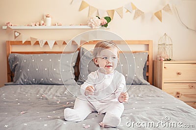 Caucasian blonde baby girl in white onesie sitting on bed in bedroom Stock Photo