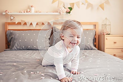 Caucasian blonde baby girl in white onesie sitting on bed in bedroom Stock Photo