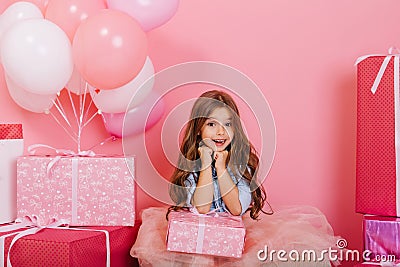 Portrait cute birthday kid expressing to camera suround big giftboxes, balloons on pink background. Amazing Stock Photo