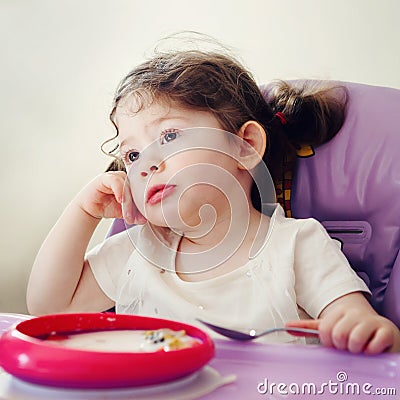 Portrait of cute bored Caucasian child kid girl sitting in high chair eating cereal with spoon early morning Stock Photo