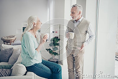 Portrait of cute charming married people in denim jeans trousers teal brown sweaters holding mugs sitting on couch in Stock Photo