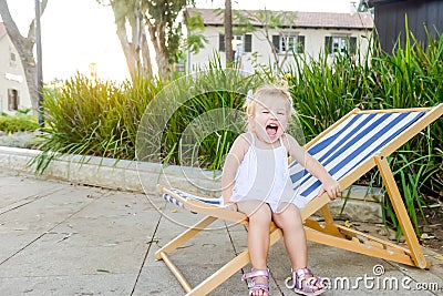 Portrait of cute emotional blondy toddler girl in white dress sitting on the deckchair and yelling. City park recreation area. Sel Stock Photo