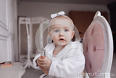 Portrait of A cute little girl in a bathrobe is doing make up and looking in the camera at home. baby Girl is sitting on the chair Stock Photo