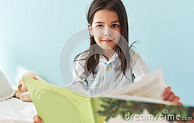 Portrait of a cute little girl sitting in the bed and reading the fairy tale book. Happy child in pajamas reading interesting Stock Photo