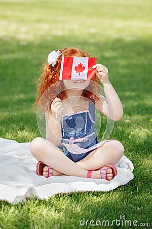 Portrait of cute little red-haired Caucasian girl child holding Canadian flag with red maple leaf, sitting on grass in park outsi Stock Photo