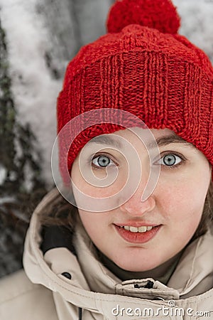 Portrait of cute thoughtfully girl with big blue eyes. Smiling young woman in red hat stands near snow-covered tree Stock Photo