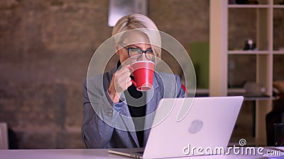 Portrait de femme d'affaires aux cheveux courts blonde en verres fonctionnant attentivement avec l'ordinateur portable et le café banque de vidéos Portrait de femme d'affaires aux cheveux courts blonde en verres fonctionnant attentivement avec l'ordinateur portable et le café banque de vidéos