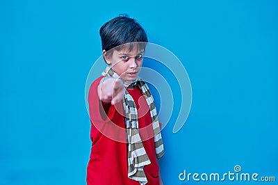 Portrait of defiant boy in red t-shirt and scarf, looking and pointing at camera with the index finger of his right hand, on blue Stock Photo