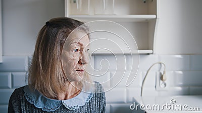 Portrait of elderly gray haired lady looking through the kitchen window and thinking. Vulnerable lonely people during Stock Photo