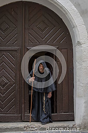 Portrait of an elderly hunched monk 45-50 years old with a beard in a black cassock with a staff coming out of the textured gate o Stock Photo